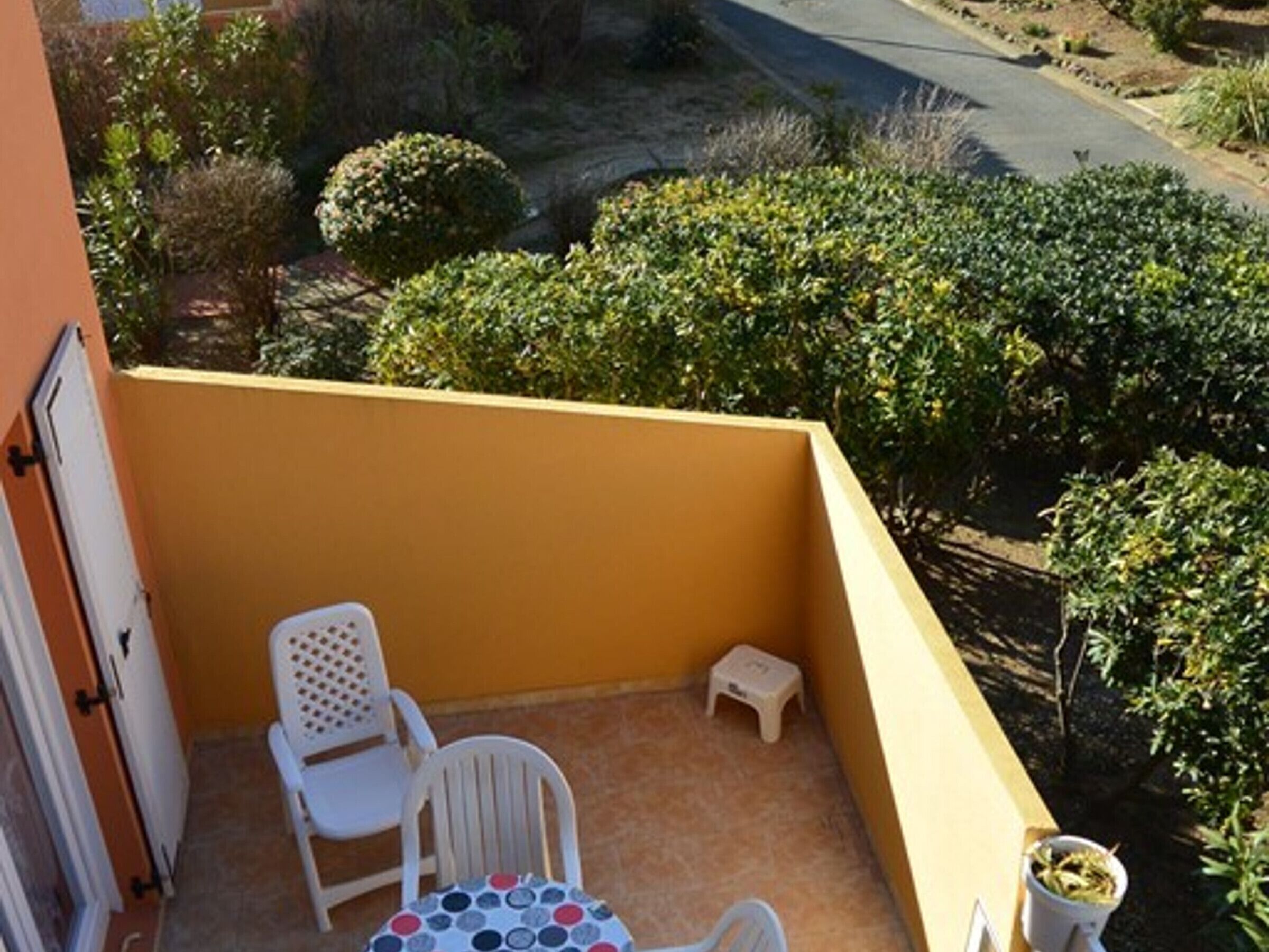 A small balcony with a yellow wall, two white chairs, a small stool, and a potted plant.