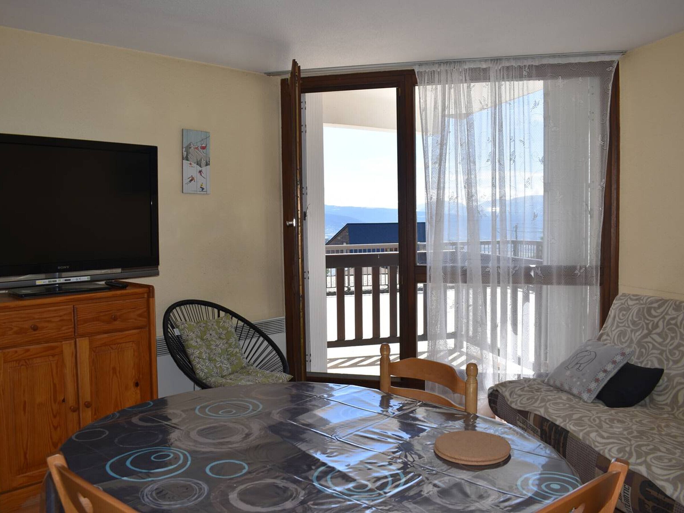 Een kamer met een televisie op een houten kast, een eettafel met stoelen, een bank en een balkon met uitzicht.