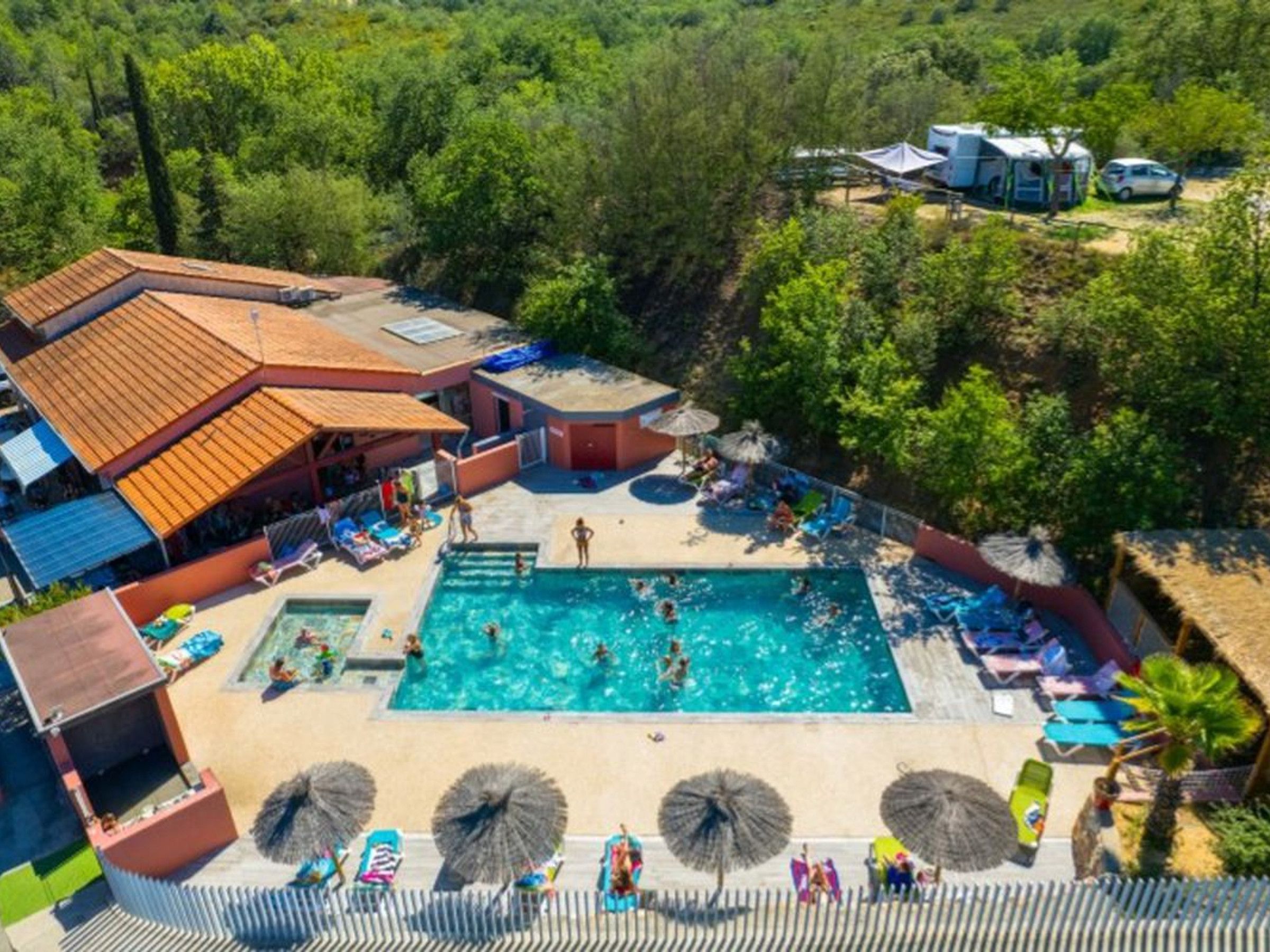 Een resort met een centraal gelegen zwembad, omringd door ligbedden en parasols, een overdekt gedeelte met een betegeld dak en een bebost achtergrond.