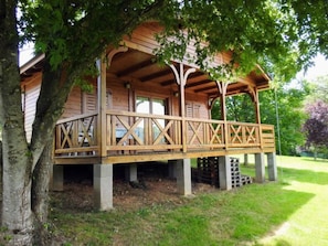 A wooden cabin with a porch supported by concrete pillars, surrounded by trees. - Cozy mobile home with terrace at Naucelle campsite (Naucelle)