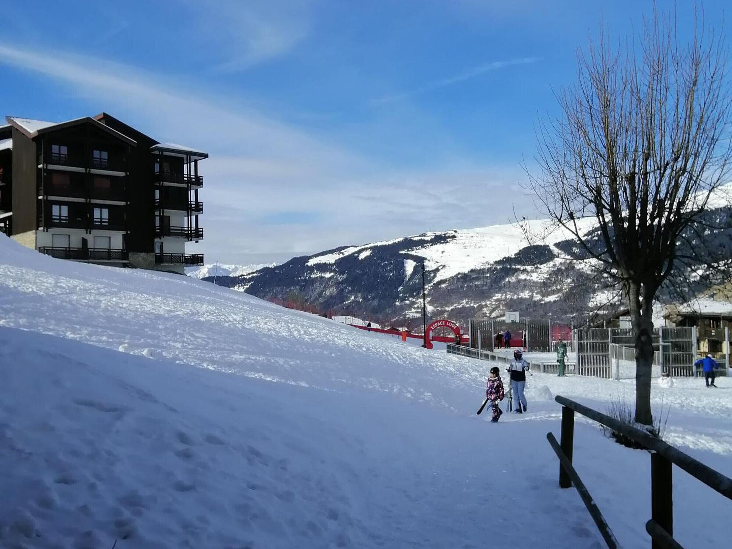A snow-covered slope with a ski lift, a building with balconies, and a mountain in the background.