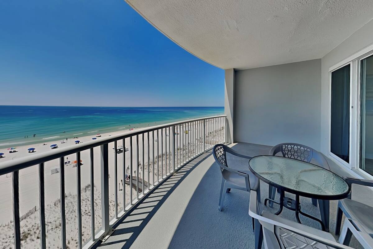 A balcony with a view of the beach and ocean, a glass table, and a chair.