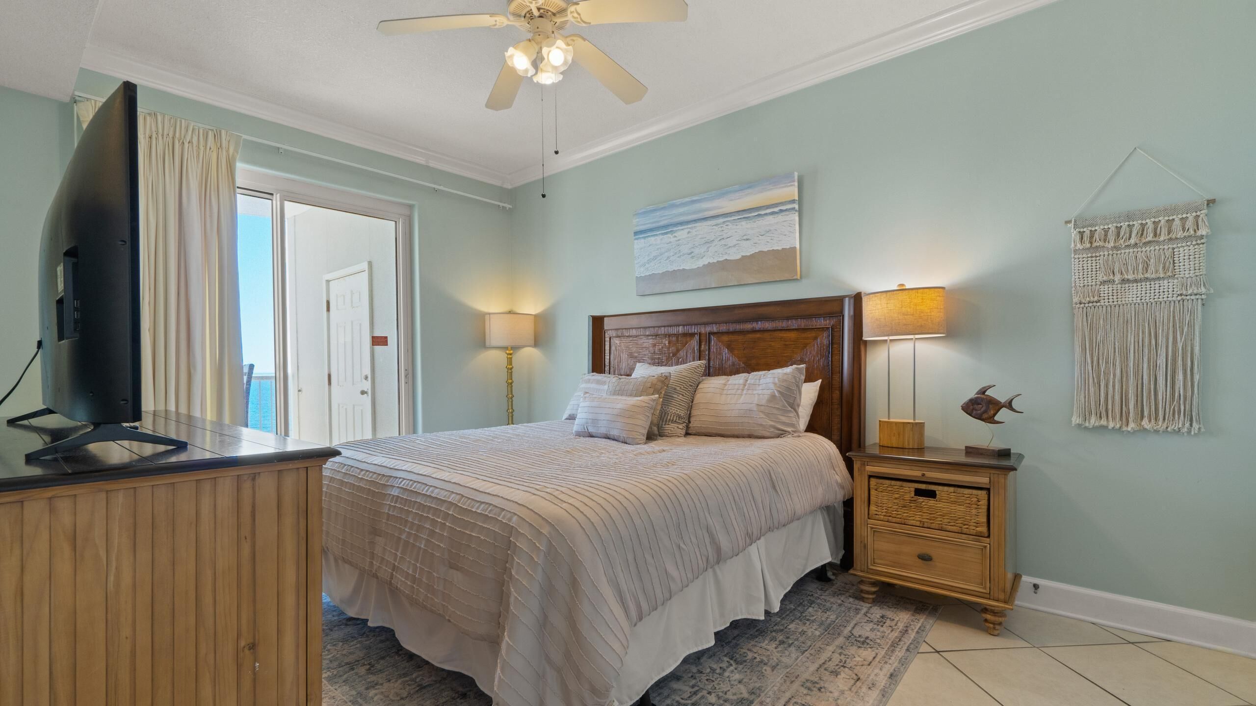 Une chambre à coucher comprenant un lit, des tables de chevet, un téléviseur, un ventilateur de plafond, un tableau et une décoration murale en macramé.