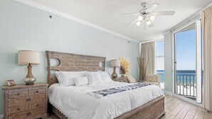 Quarto com cama grande, cabeceira de madeira, duas mesas de cabeceira com luminárias, vista para o oceano através de uma porta de varanda e um ventilador de teto.