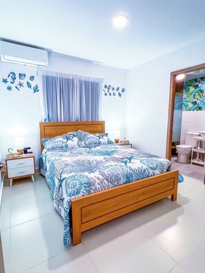 A bedroom with a wooden bed, white walls, a bathroom with a white toilet, and a shower area. - Themed & Peaceful -Garden view - Energy Included (Punta Cana)