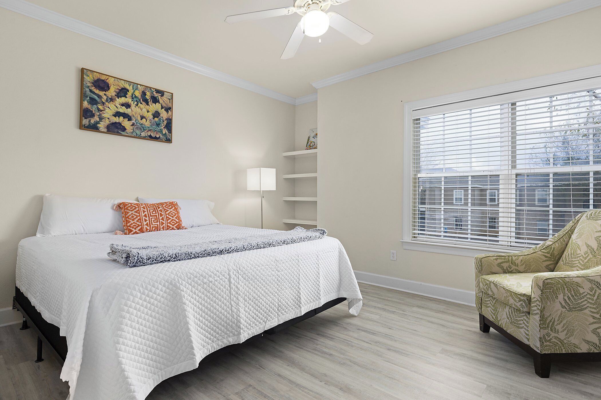 A bedroom with a bed, a chair, a ceiling fan, a window with blinds, and a painting on the wall.