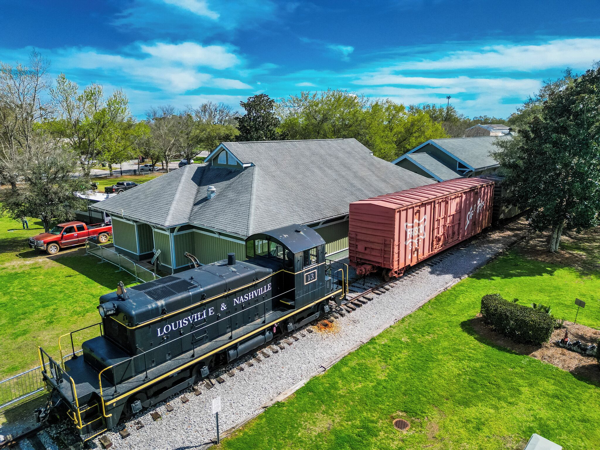 A train with "Louisville & Nashville" on the front, passing by a building with a green roof and a red truck parked nearby.