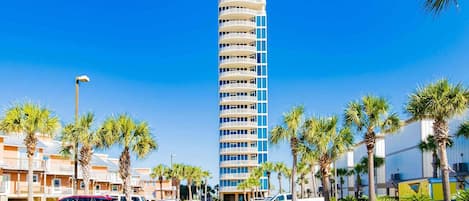 A tall, multi-story hotel with a modern design, featuring balconies on each floor, surrounded by palm trees and parked vehicles.