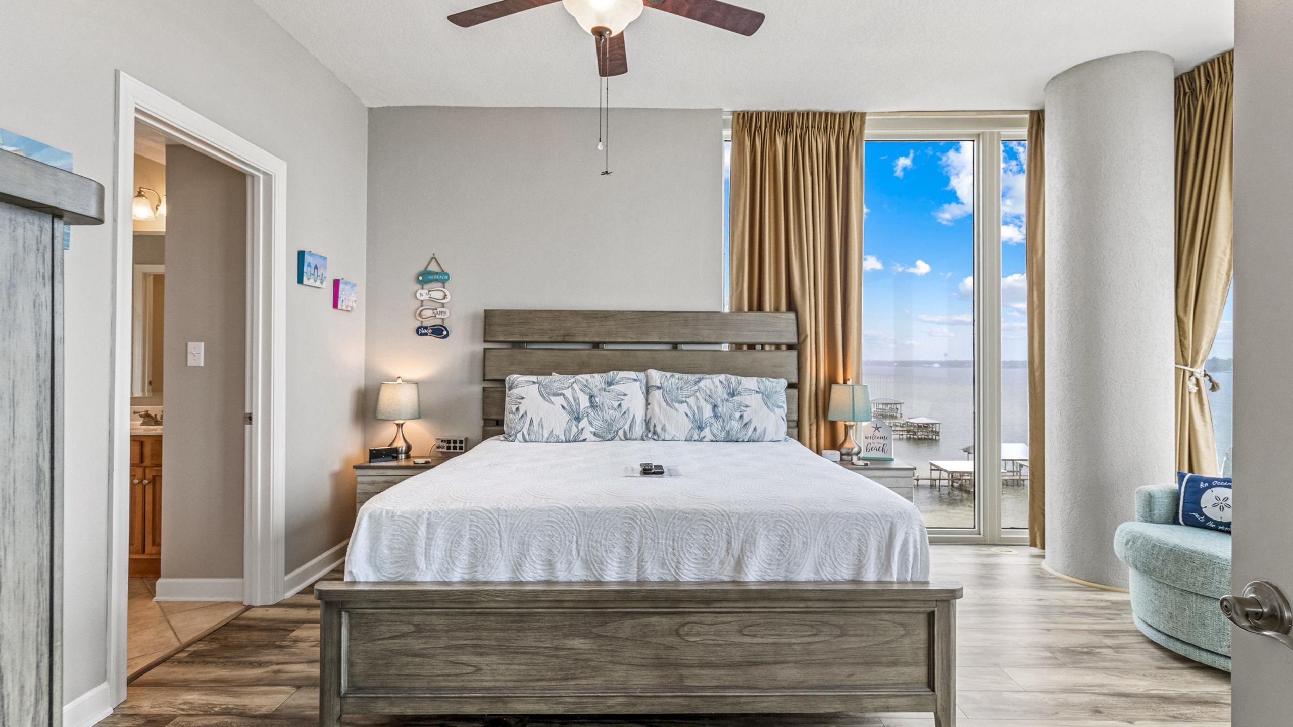 A bedroom with a wooden bed, a ceiling fan, a view of the ocean, and a balcony with outdoor seating.