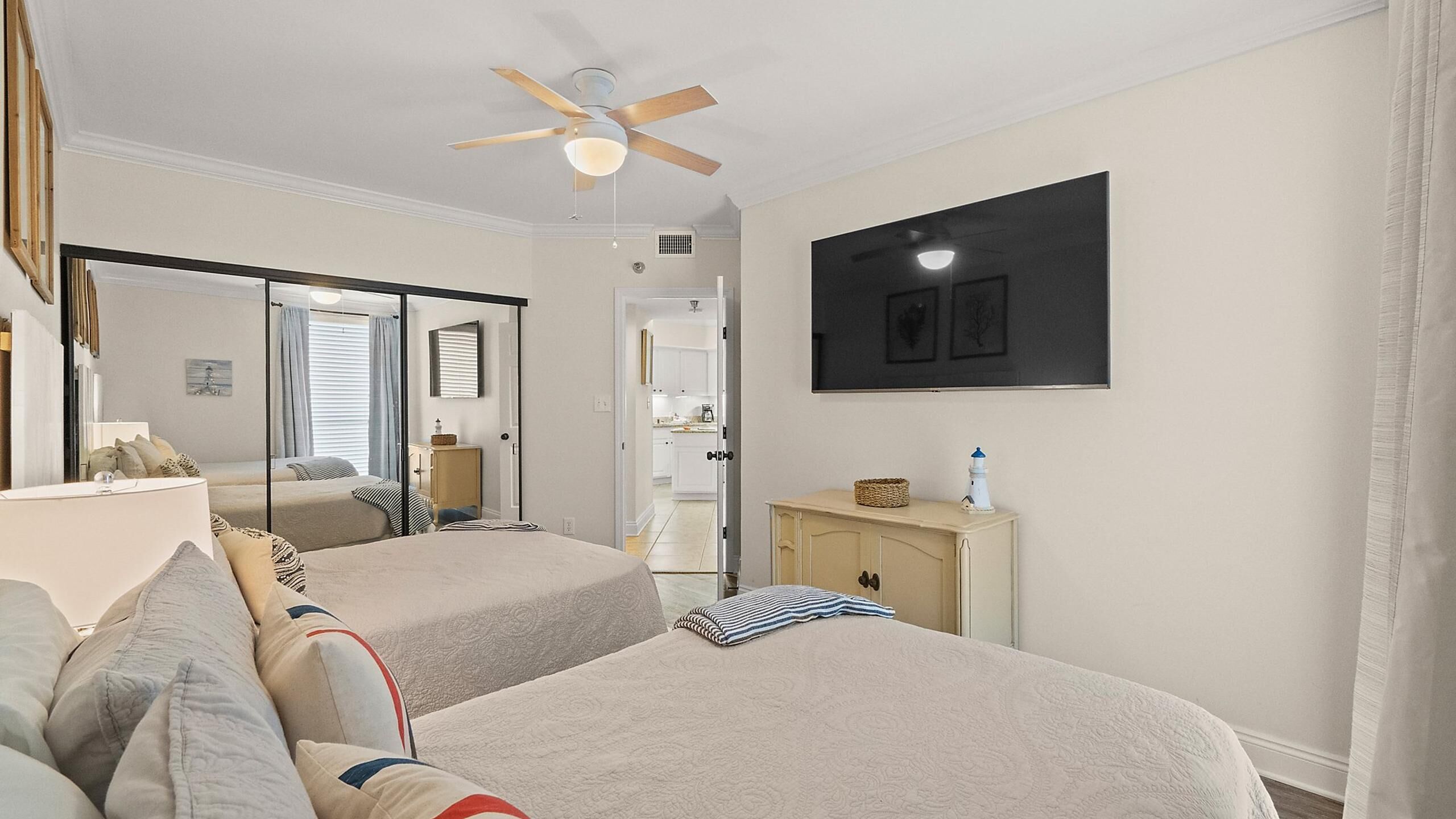 Une chambre avec deux lits, un ventilateur de plafond, un miroir, une petite armoire et un téléviseur fixé au mur.