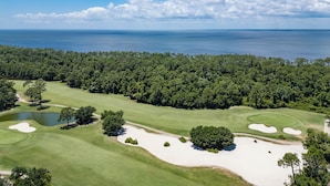 Un campo de golf con búnkeres de arena, un estanque y un bosque denso al fondo.