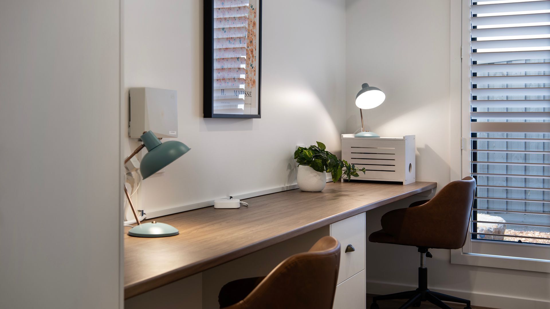 Een modern bureau met een lamp, een kamerplant en een lijst aan de muur.