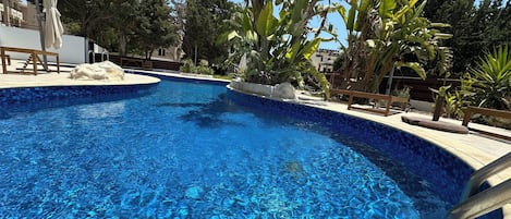 A clear blue pool with a tiled edge, surrounded by tropical plants and a wooden deck.
