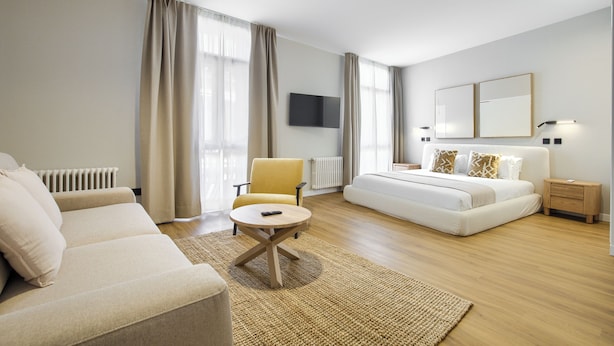 A modern hotel room with a sofa, a round coffee table, a bed with a headboard, and a flat-screen TV mounted on the wall. - Dobo Madrid Justicia (Madrid)
