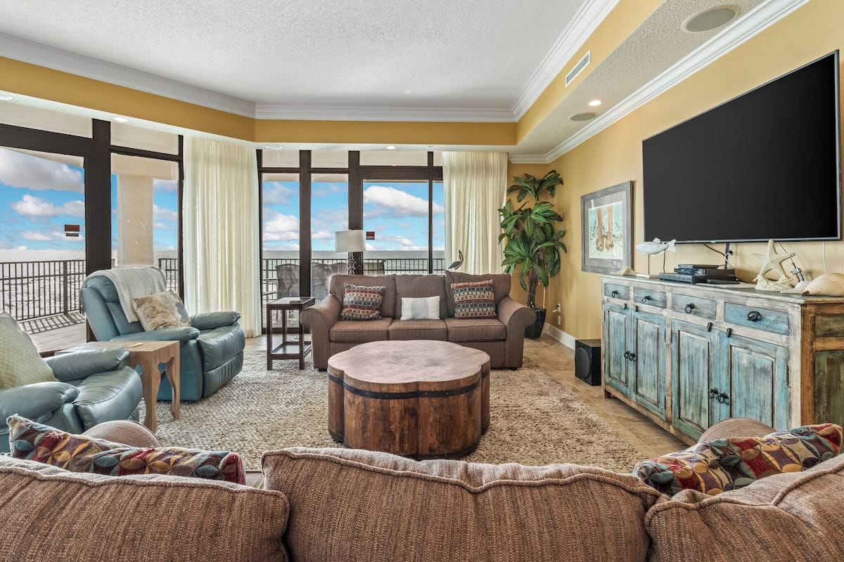 A spacious living room with a large sofa, a round coffee table, and a television. There is a balcony with a view of the sky.
