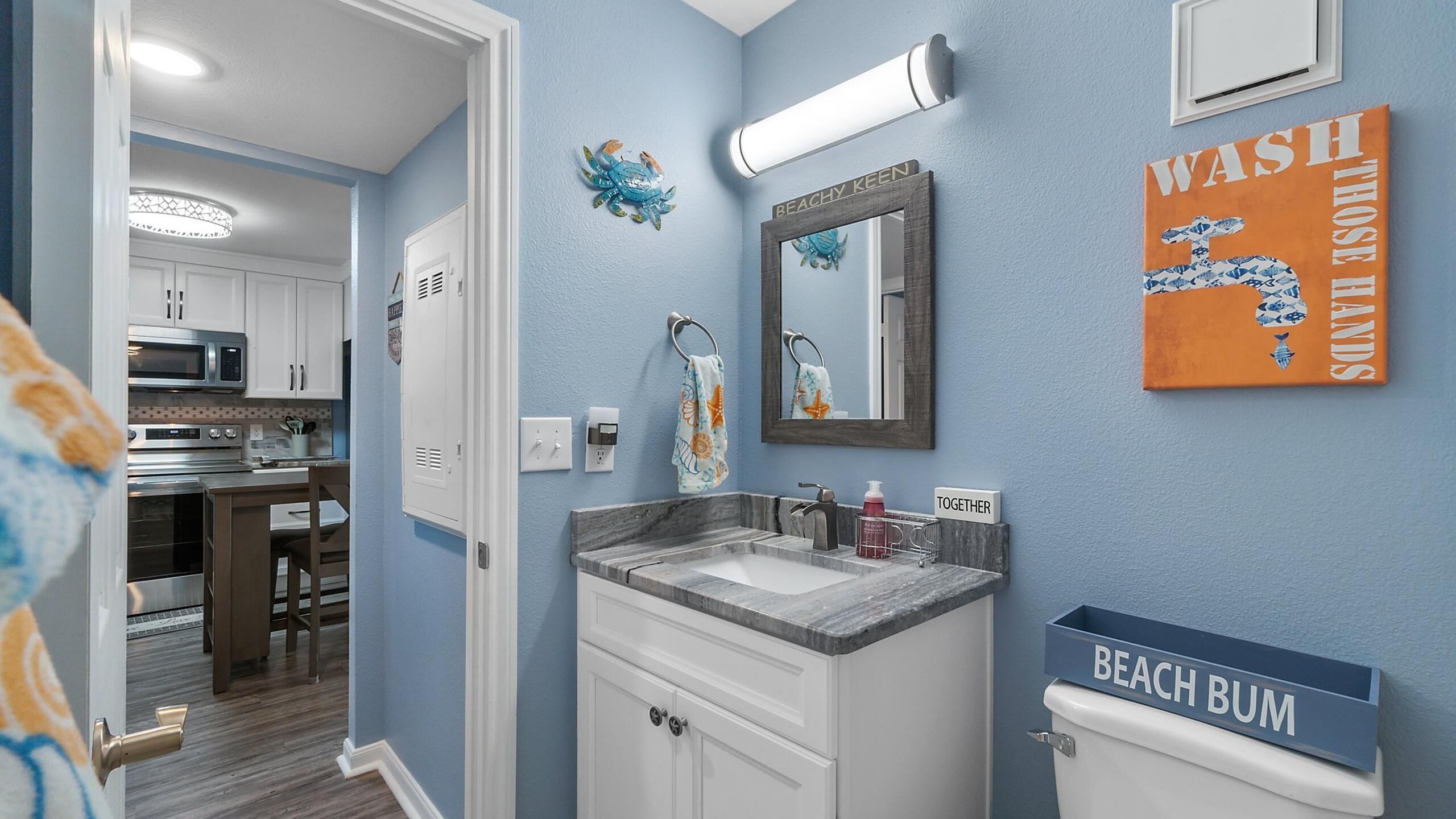 A bathroom with a sink, mirror, and towel rack. There is a sign that says "BEACH BUM" and another that says "TOGETHER".