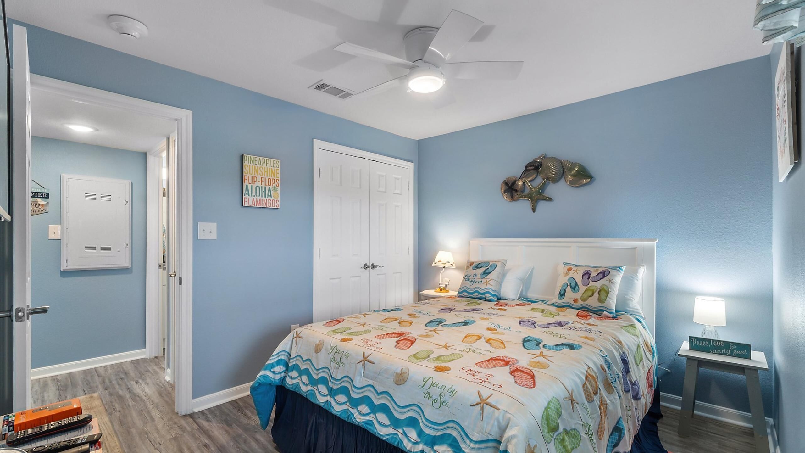 Une chambre à coucher avec un lit recouvert d’une couvre-lit sur le thème de la plage, un ventilateur de plafond et un mur décoré de coquillages et d’une étoile de mer.