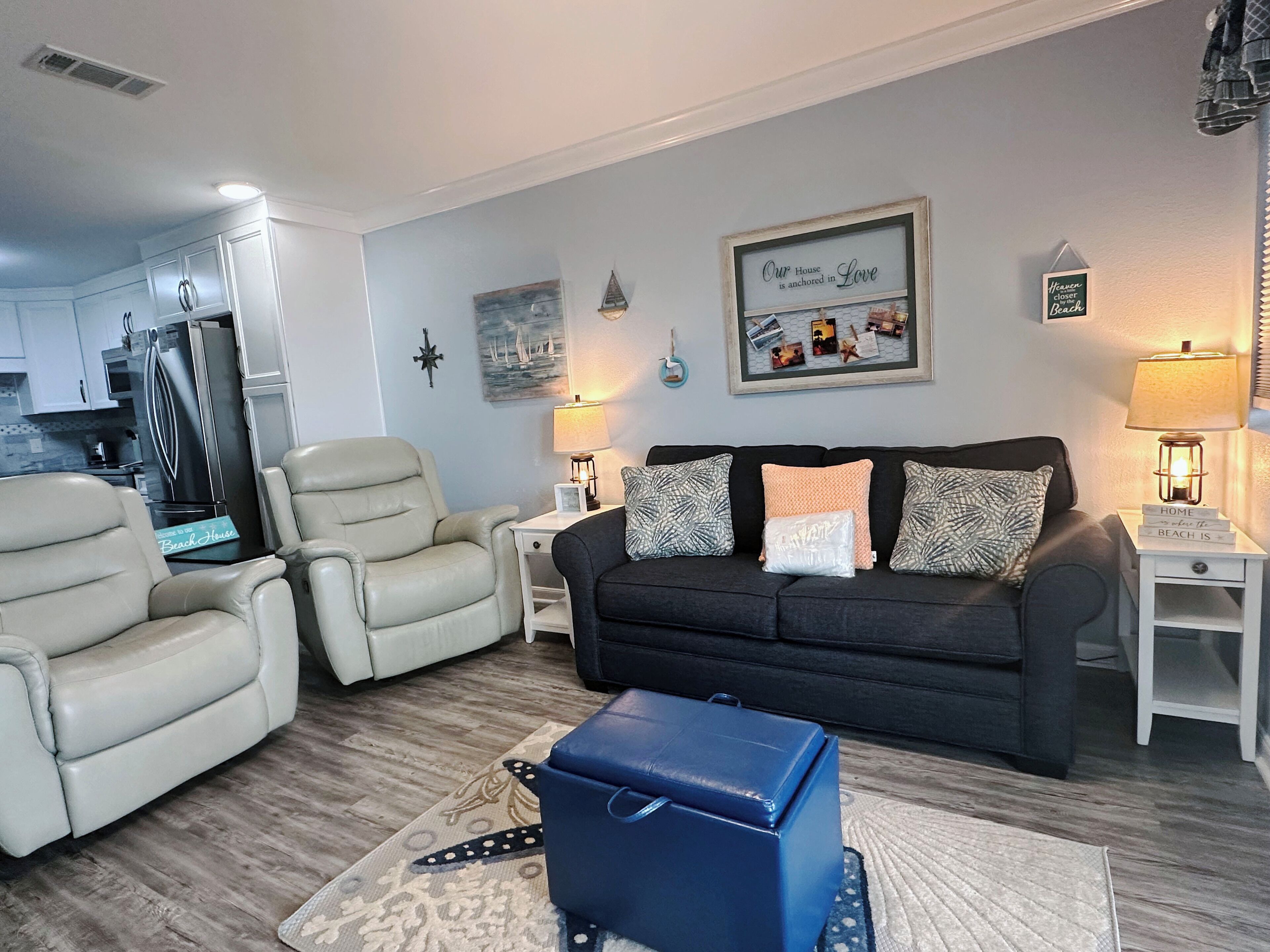 A living room with a sofa, two armchairs, a coffee table, and a small blue ottoman. There is a kitchen in the background with a refrigerator and a stove.