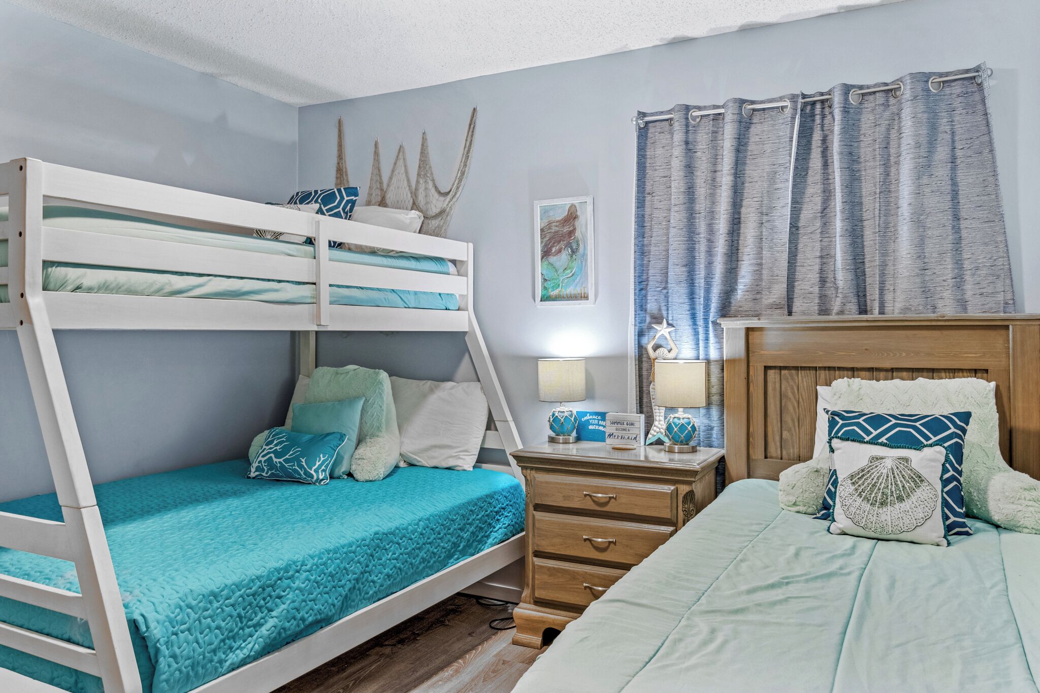 A bunk bed with a teal bedspread, a wooden nightstand with a lamp, and a headboard with decorative pillows.