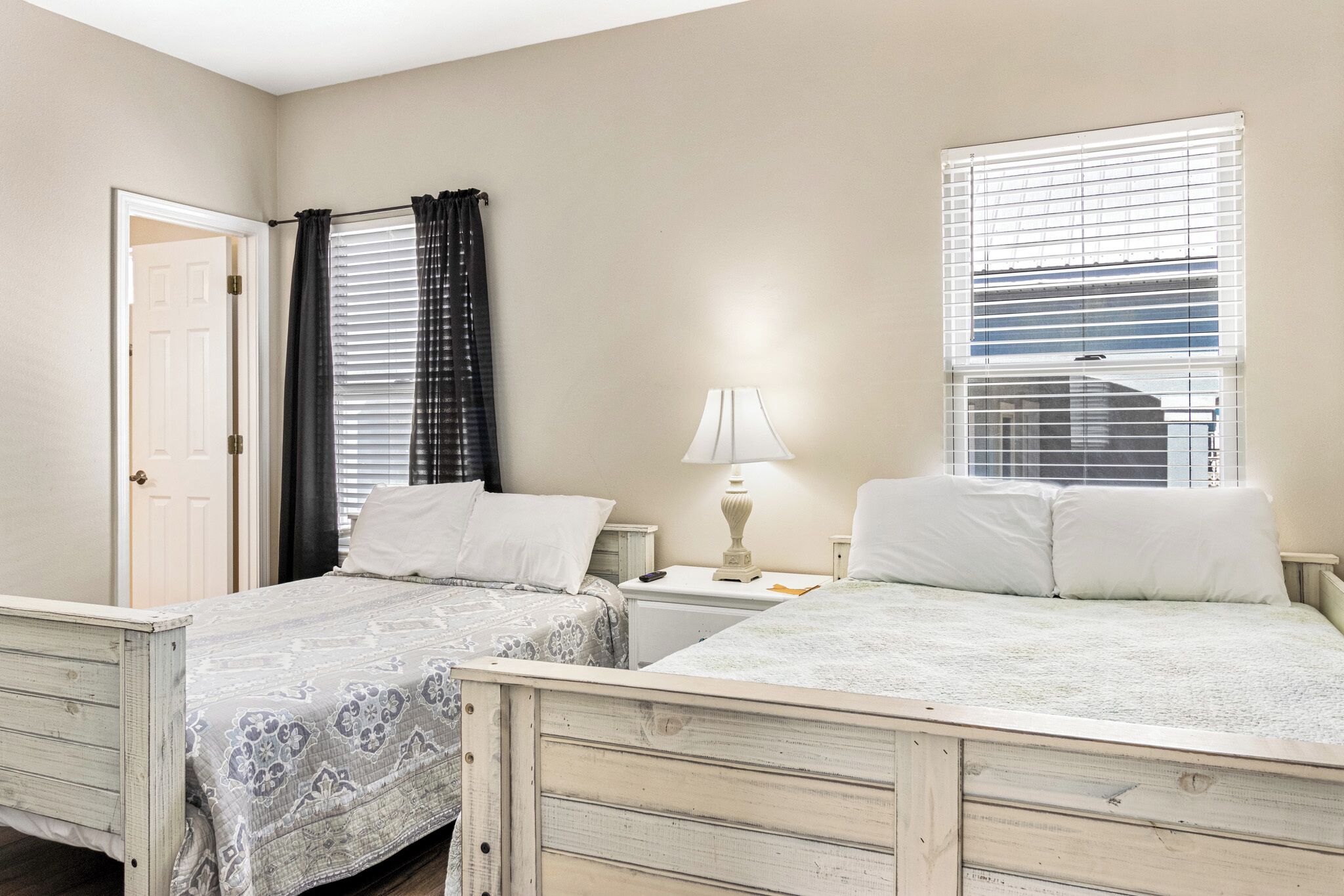 A bedroom with two beds, a door, a window with blinds, and a nightstand with a lamp.