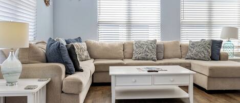 A living room with a beige sectional sofa, white coffee table, and a small side table with a lamp. There are two windows with blinds, a wall-mounted ship's wheel, and a paddle on the wall.