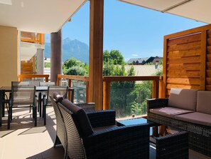 Un balcon ensoleillé, agrémenté de meubles en osier, d’une table en verre et offrant une vue sur les montagnes.