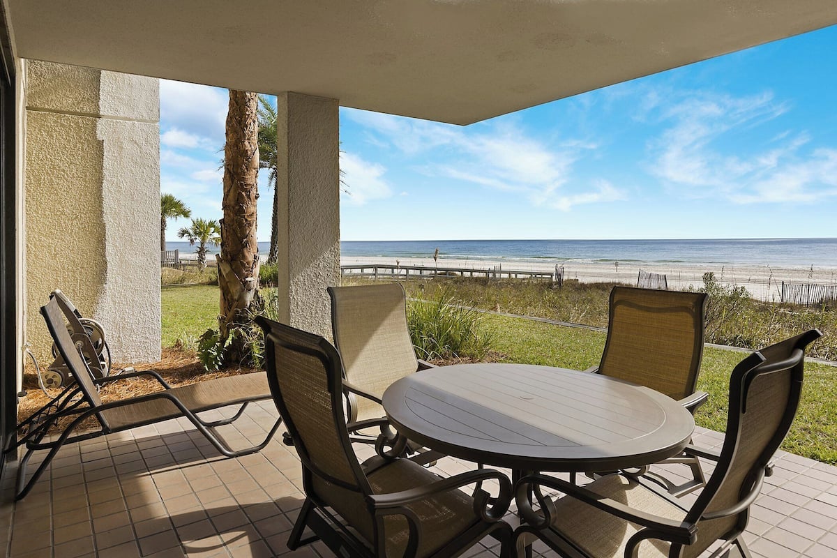 Een terras met een ronde tafel en stoelen, met uitzicht op een strand en de oceaan.