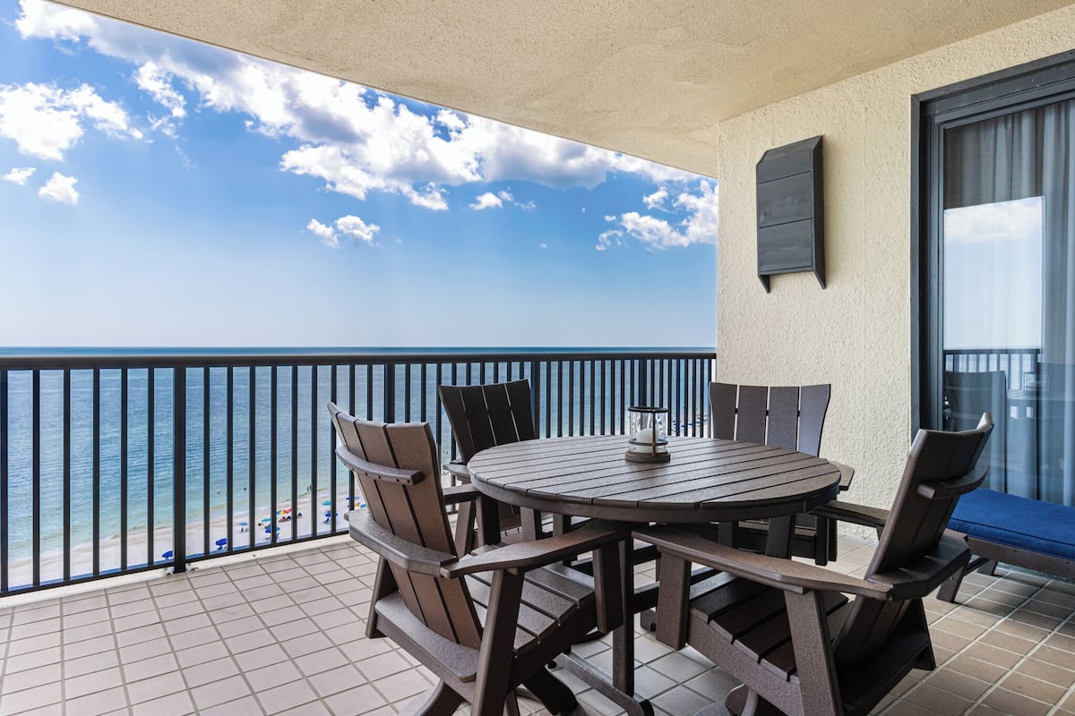 Een balkon met een ronde tafel en stoelen, met uitzicht op een strand met parasols en een heldere lucht.