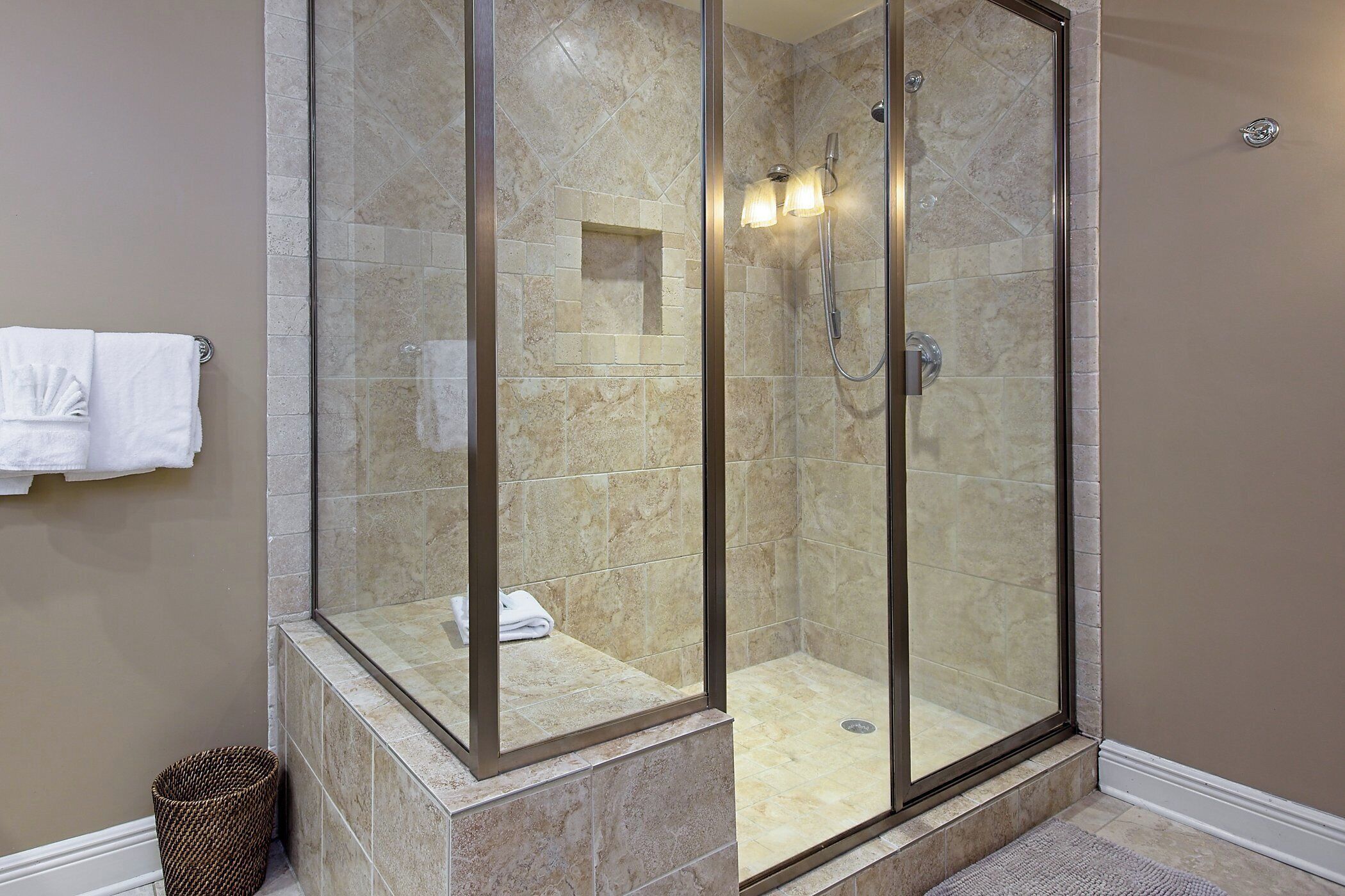 A glass-enclosed shower with a tiled floor and a small shelf, a towel rack with a towel, and a waste basket.