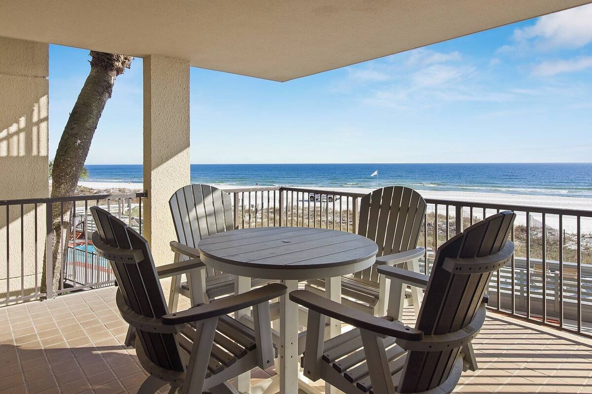 Een balkon met een tafel en stoelen, uitkijkend op een strand met een helder uitzicht op de oceaan.