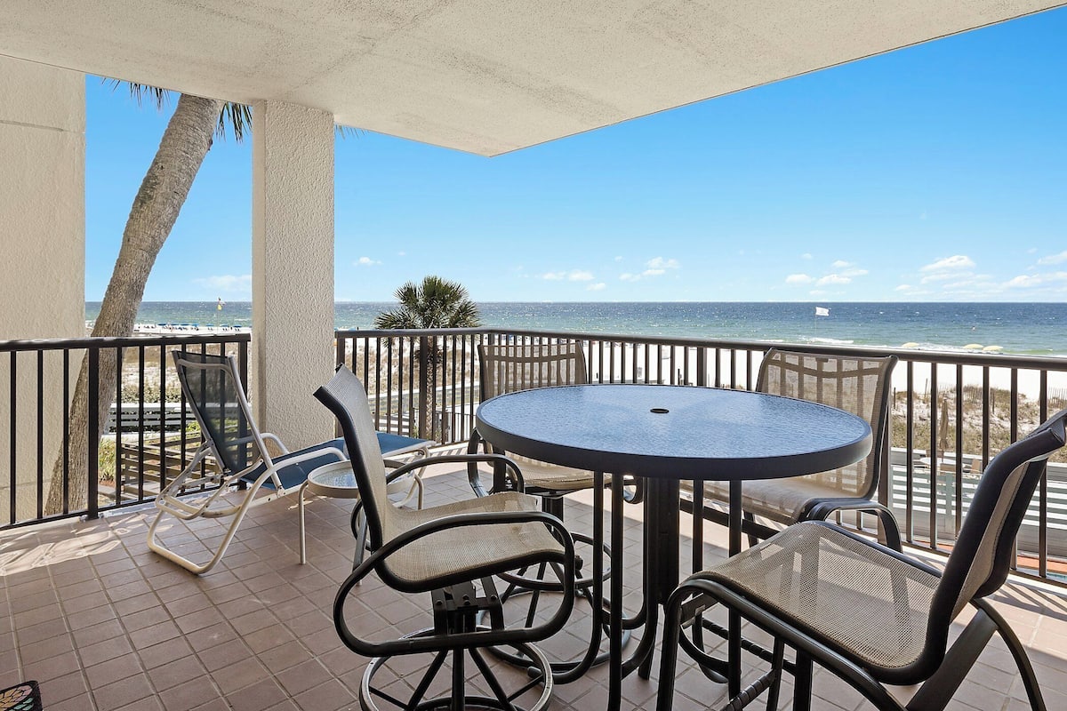 Een balkon met een ronde tafel en stoelen, met uitzicht op een strand en een helder zicht op de oceaan.