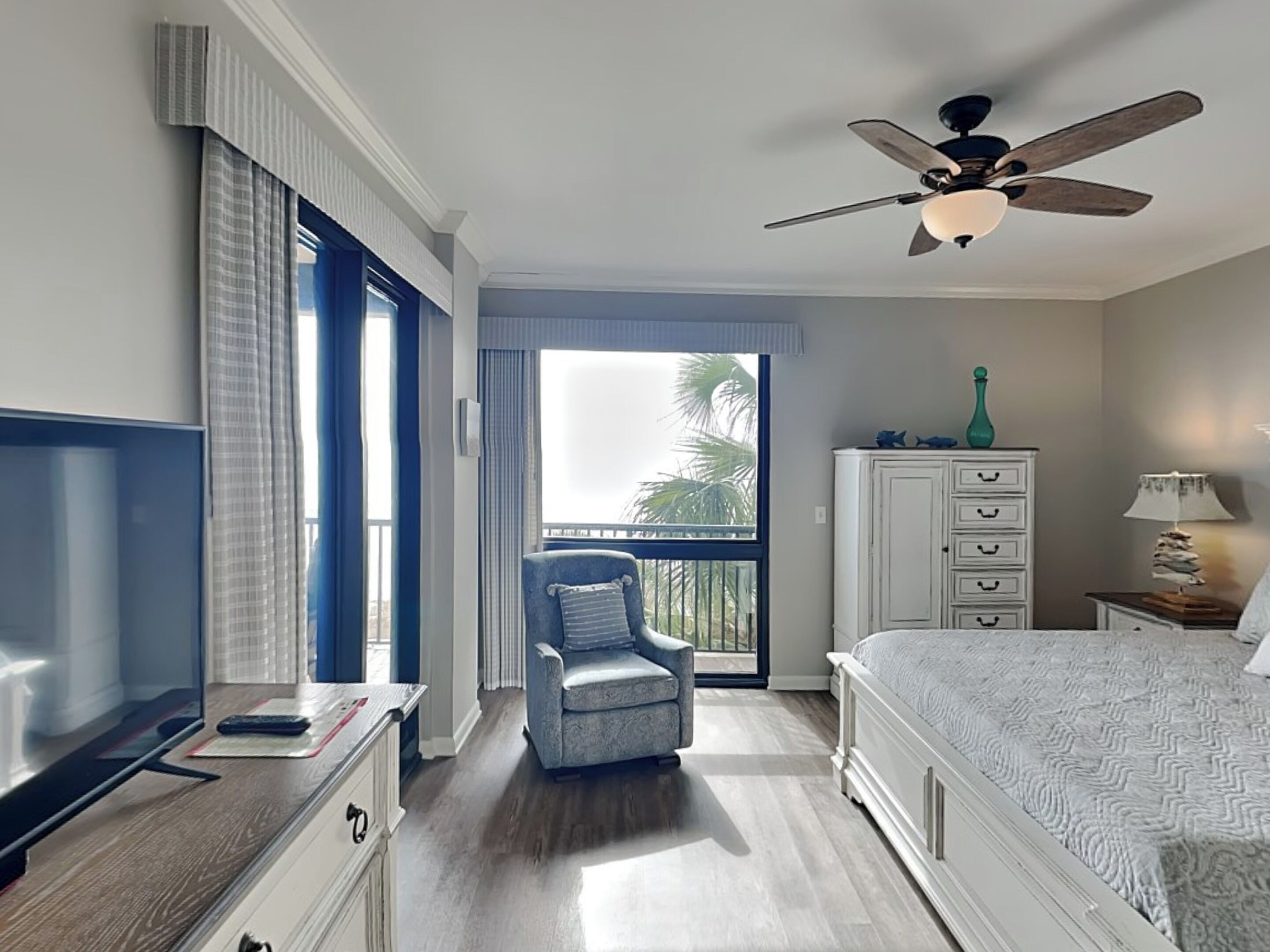 Et hotelværelse med seng, stol, skrivebord med fjernsyn, garderobeskab, natbord med lampe og en ventilator i loftet.