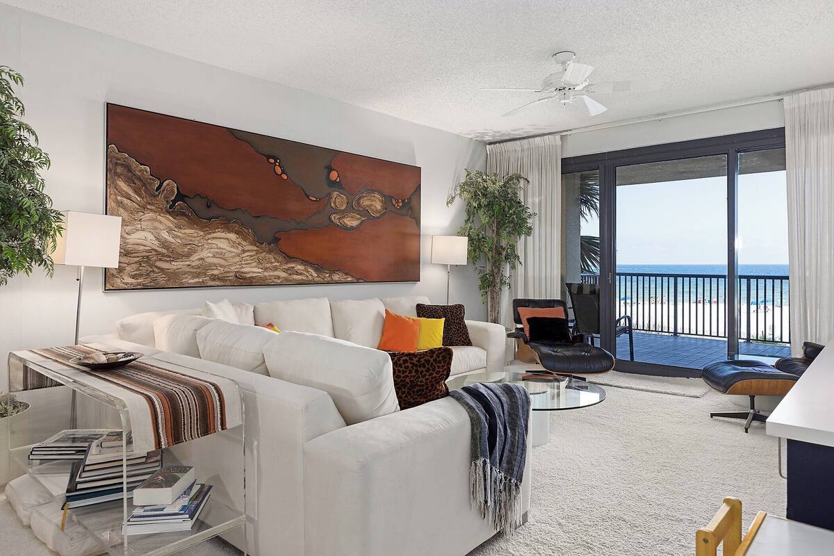 Een moderne woonkamer met een witte bank, een glazen salontafel en een groot abstract schilderij aan de muur. Er is een balkon met uitzicht op de oceaan.