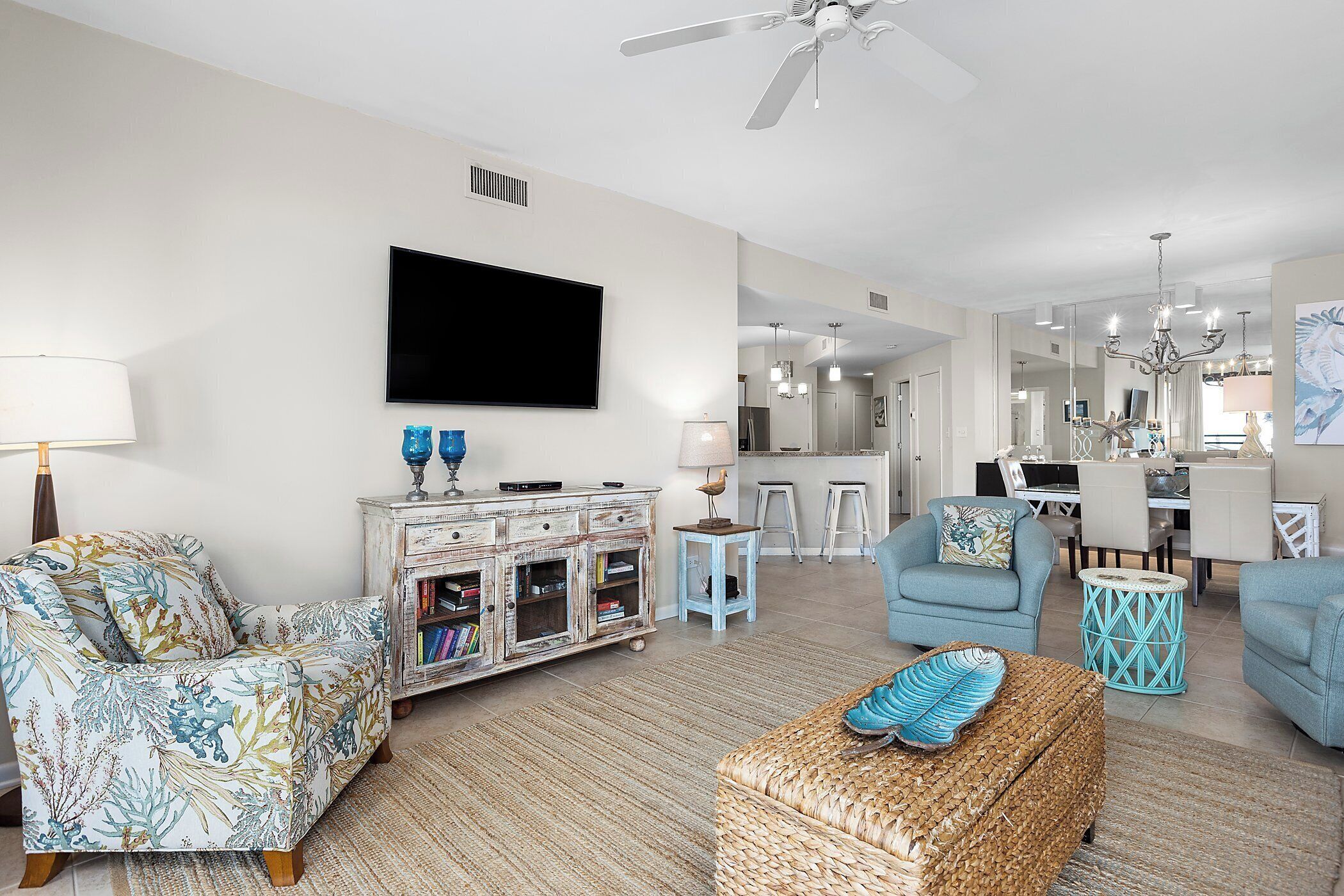 A living room with a flat-screen TV, a coffee table, and a sofa. There is a dining area with a chandelier and a kitchen in the background.