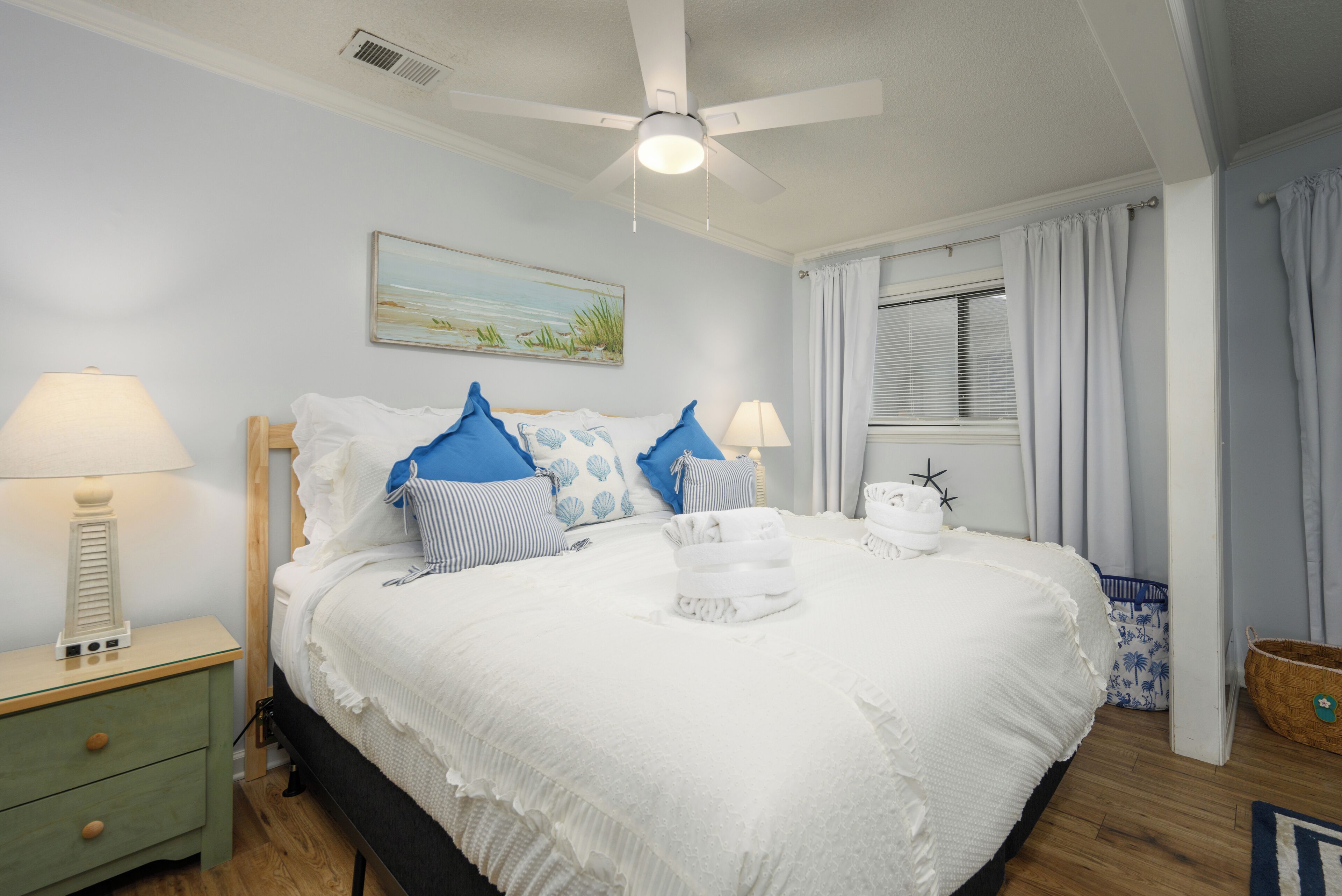 Ein ordentlich bezogenes Bett mit weißer Decke und blauen Kissen, ein Deckenventilator, ein Nachttisch mit Lampe und ein Fenster mit Vorhängen.