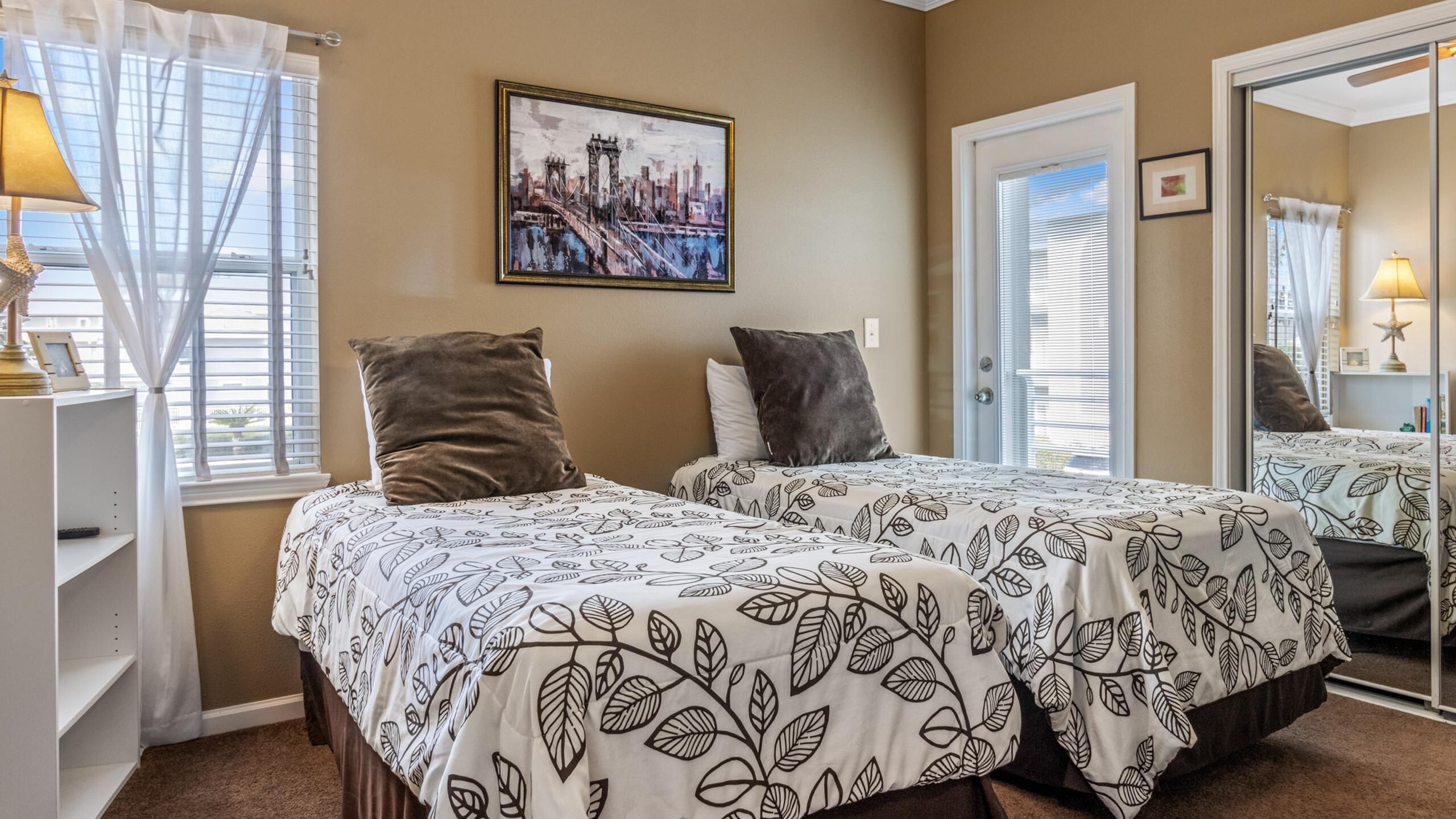 A hotel room with two beds, a large mirror, a framed cityscape painting, and a window with curtains.
