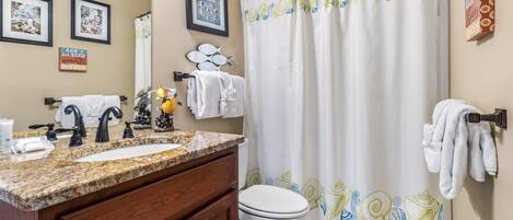 A bathroom with a granite countertop sink, two black faucets, a white toilet, and a shower curtain with a shell pattern.