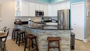 A kitchen with white cabinets, a stainless steel refrigerator, a microwave, and a stone island with bar stools. - San Carlos #605 by Southern Vacation Rentals (Gulf Shores)
