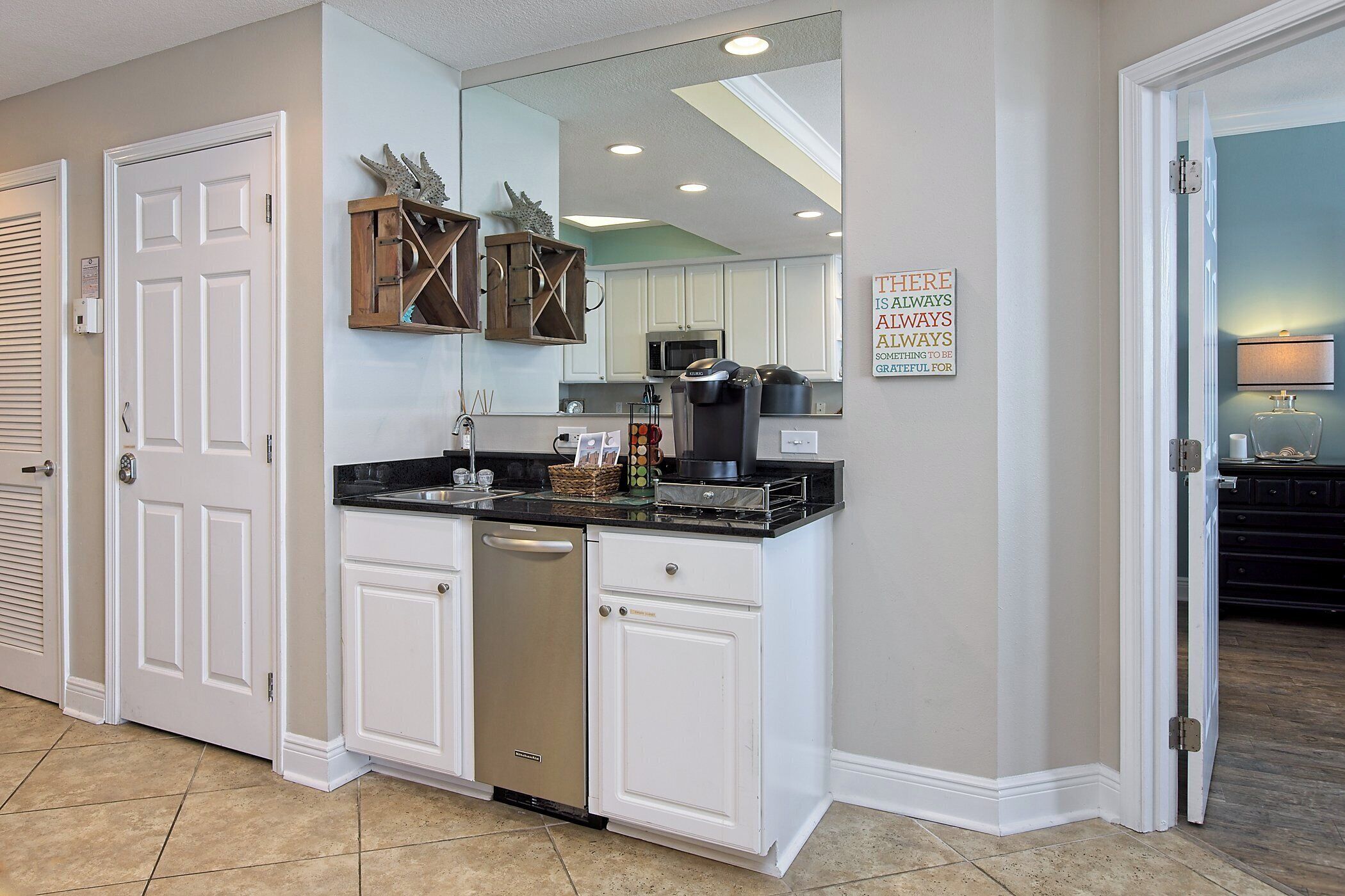 A kitchen with white cabinets, a dishwasher, and a microwave. There is a sign on the wall that says "HERE IS ALWAYS SOMETHING OF GREATFUL FOR."