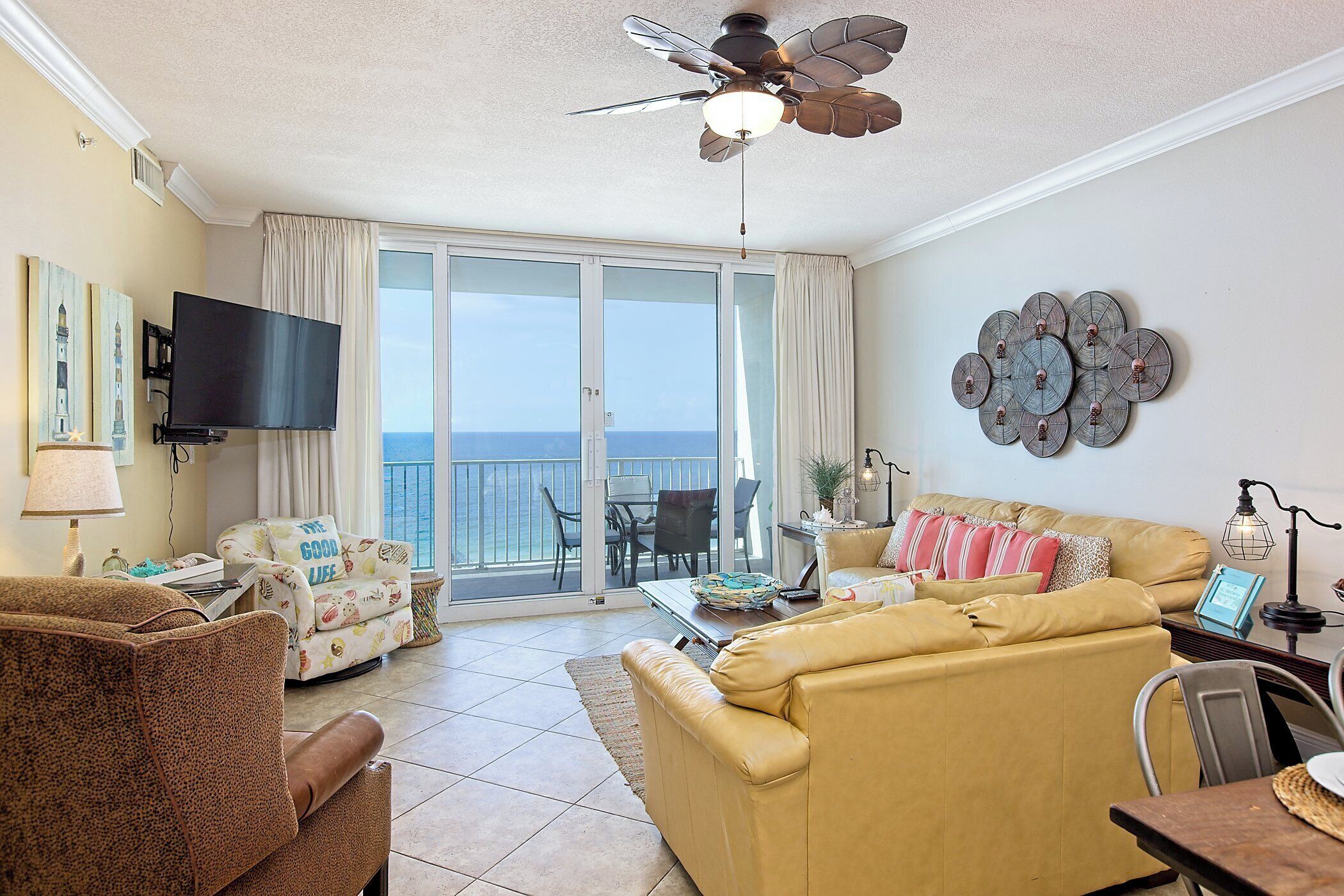 A living room with a sofa, armchair, and a television. There is a balcony with a view of the ocean.