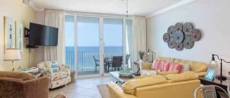 A living room with a sofa, armchair, and a television. There is a balcony with a view of the ocean.