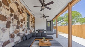 A covered patio with a stone wall, a sectional sofa, a wooden coffee table, and a ceiling fan.