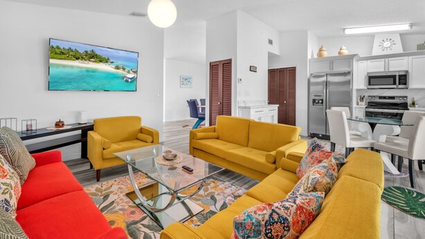 Ein modernes Wohnzimmer mit einem großen Flachbildfernseher, gelben Sofas, einem Glastisch und einer Küchenzeile mit weißen Schränken und Geräten.