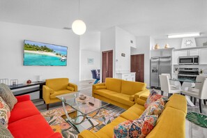 A modern living room with a large flat-screen TV, yellow sofas, a glass coffee table, and a kitchen area with white cabinets and appliances. - Lime Haus 🍋 | Zesty 3BR Escape with Private Yard (Miami)