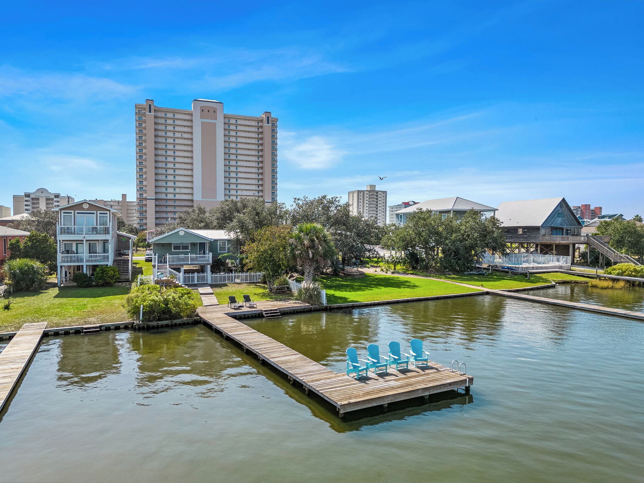 A waterfront hotel with a dock and chairs, surrounded by greenery and a large building in the background.
