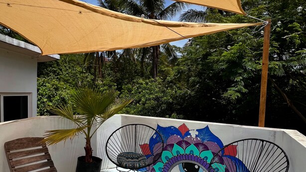 Une terrasse sur le toit avec une chaise en bois, deux chaises en fil de fer, une plante en pot et une fresque murale colorée.