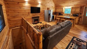 A log cabin interior with a fireplace, a sofa, a kitchen area, and a television mounted on the wall.