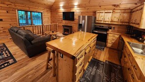 A log cabin interior with a sofa, fireplace, kitchen area, and a mounted deer head.