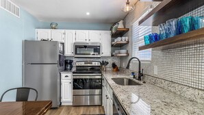 A compact kitchen with white cabinets, stainless steel appliances, a granite countertop, and a blue rug. - Sunrise Village #210 by Southern Vacation Rentals (Gulf Shores)