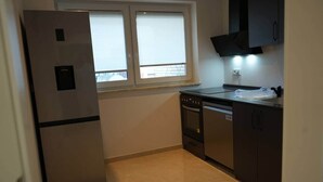 Cocina compacta con refrigerador, horno y armarios, una ventana con persianas, y piso de baldosas.